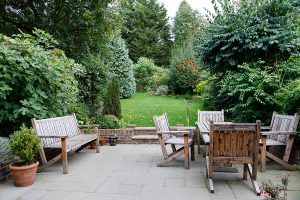 Landscaped backyard with green trees and bushes with a large patio with natural wood furniture. 